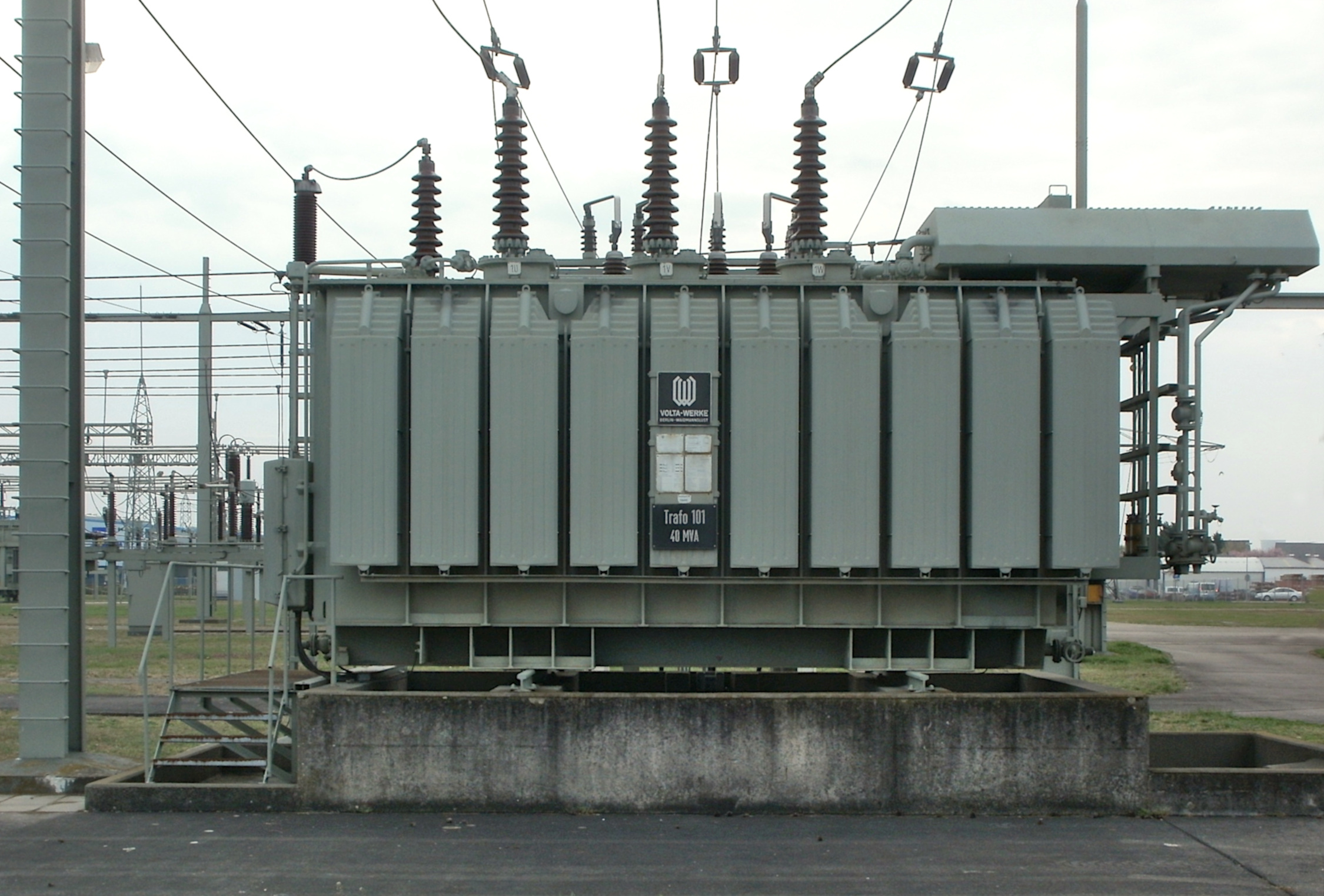 Ein großer, rechteckiger Leistungstransformator in grauer Farbe steht in einer Freiluftanlage. Er hat
mehrere Hochspannungsleitungen, die nach oben führen, und ist auf einer betonierten Fläche
platziert.